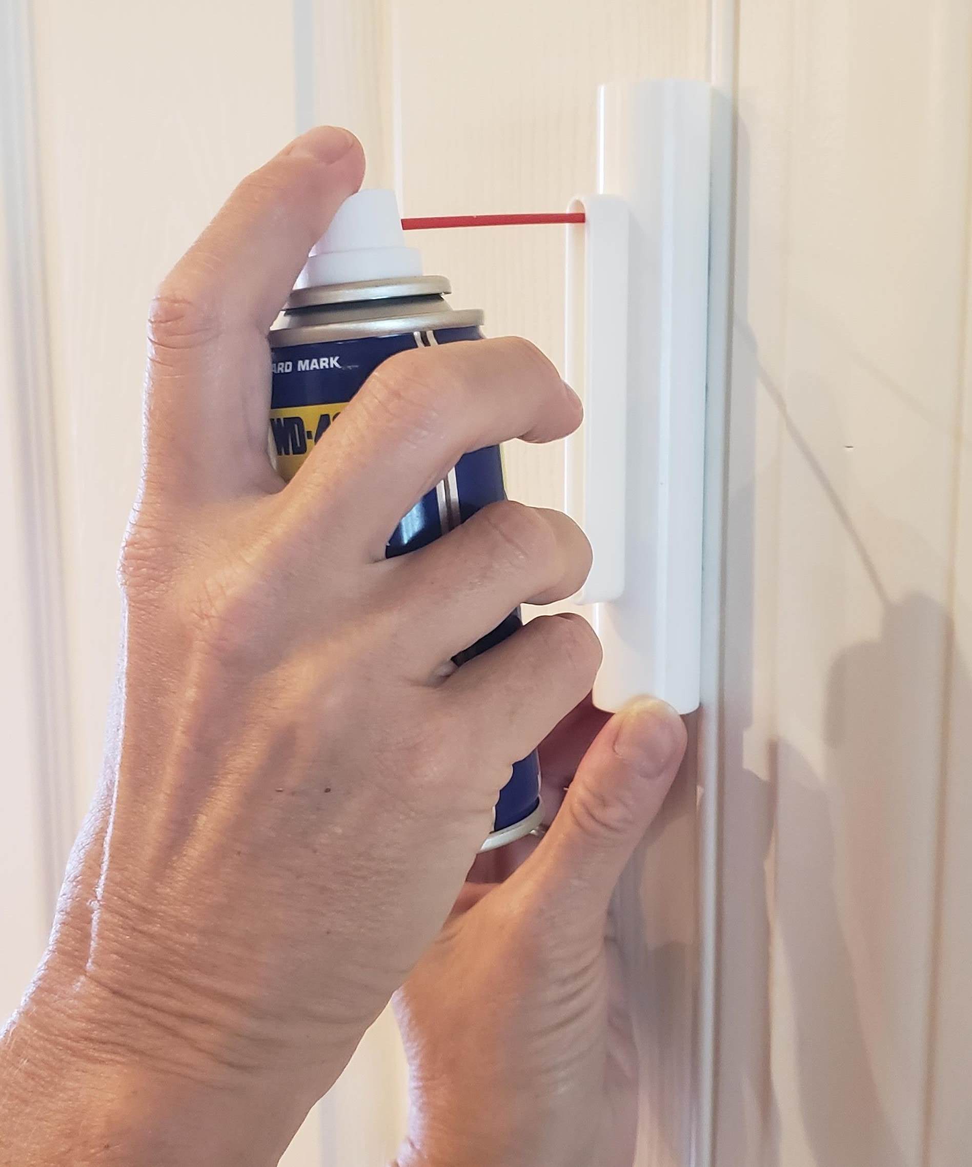 Hand using the Hinge Hero tool to apply WD-40 lubricant to a door hinge for smooth operation.