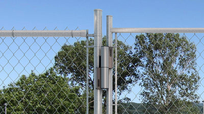 Locinox Tiger hydraulic self-closing gate closer and hinge kit installed on a chain-link fence with trees in the background.