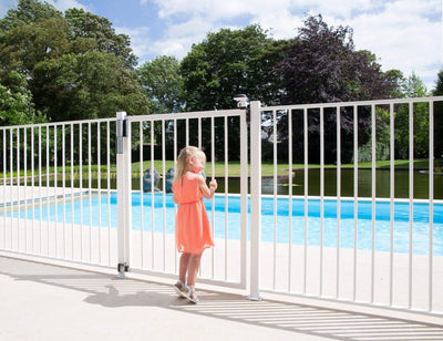 Child in a pink dress stands by a self-closing gate near a pool, showcasing the Locinox Tiger gate closer and hinge kit.