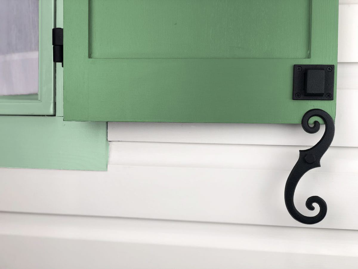 Beveled scroll shutter dogs in black powder coat, 6-3/4 inches, securing a green shutter to a white wall. Sold in pairs.