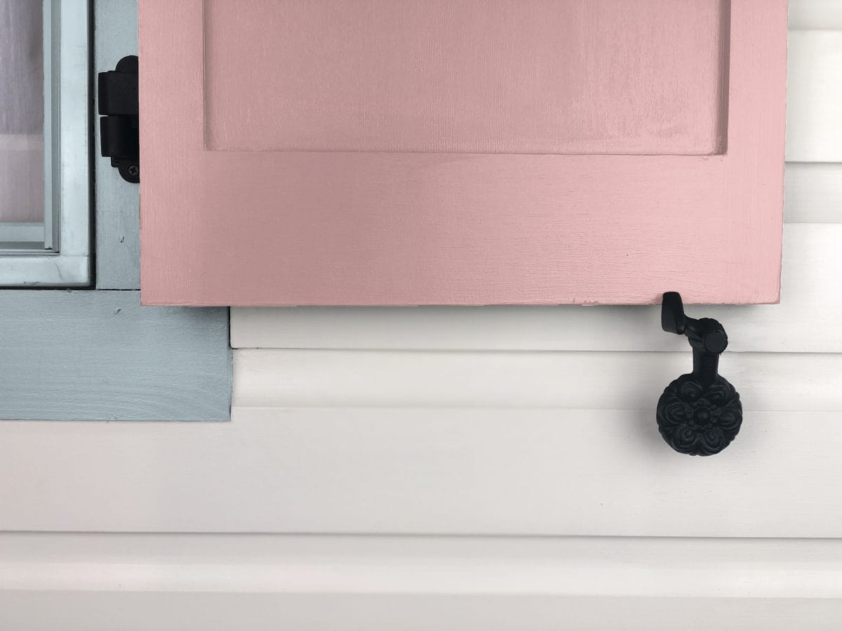 Black cast iron shutter dog tieback in floral design, securing a pink window shutter against a white wall.