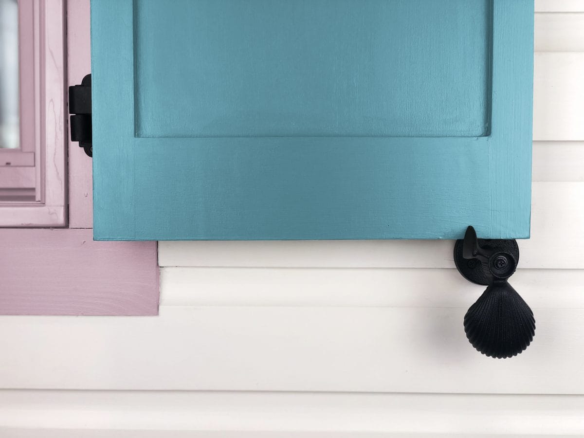 Black powder-coated cast iron shutter dogs in shell design, 5-1/4 inches, securing a blue shutter to a white wall.